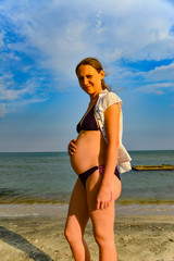 Fototapeta na wymiar A pregnant woman in a white blouse is standing on the beach.