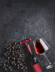 Red wine bottle with vintage corkscrew, glass and grapes on retro black background, top view.