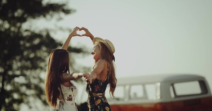 Young two women from 90 s in the middle of nature with their retro bus , making a beating heart in the air with hands.