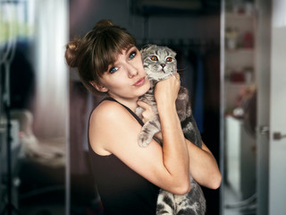 Beautiful young blonde woman in black t-shirt with high buns hairstyle looking with easy attractive smile petting scottish fold cat indoors in her room