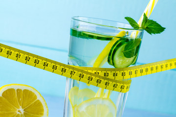Diet concept. Detox drinks. Lemon water in a glass, juicy lemon, mint and measuring tape on a blue...