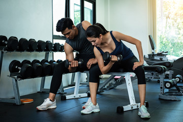 The beauty lady and handsome man are wearing exercise suit,raising dumbbell for built arm muscle,at fitness room,blurry light around