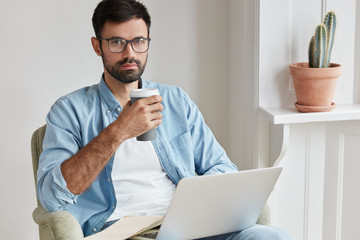 Bearded young man manager checks necessary information on laptop computer, works freelance, enjoys cappuccino. Designer studies sketches and ponders on new strategy. People, technology, work concept