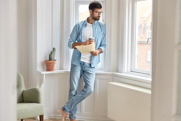 Indoor shot of porfessional marketer considers schedule productivity on papers, dressed in casual denim clothes, drinks fresh beverage from disposable cup, examines paperwork durig day off at home