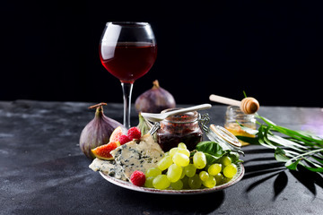 delicacy blue cheeses, fruit and jam in jar with red wine in glass and honey, palm leaf on black slate stone background, copy space