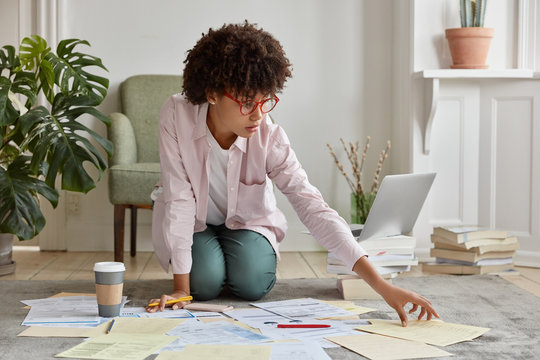 Black marketing director faces financial disclosure, studies economic forces, surrounded with much papers, dressed in casual shirt, spectacles, poses on floor of cozy apartment. Business education