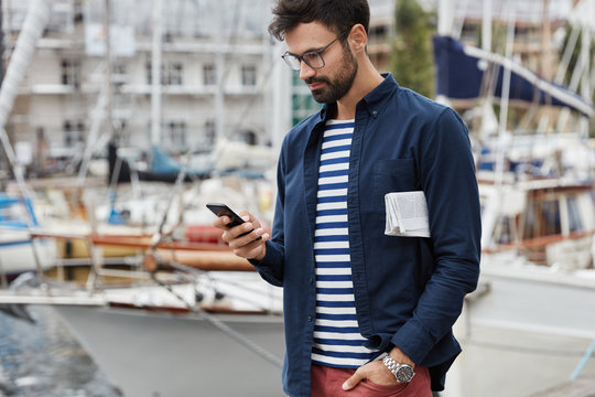 Cropped shot of hipster guy in fashionable clothes, carries newspaper, has stroll across harbour in resort place, checks email via smart phone, enjoys free internet connection. Blogger browses website