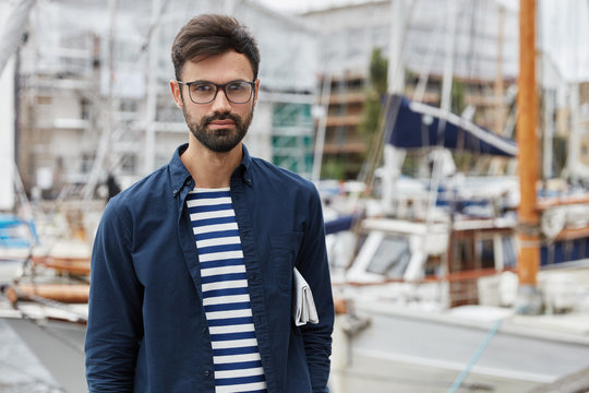 Outdoor shot of bearded young sailor dressed in fashionable clothes, has rest after arrival, poses against sailboats blurred background, carries newspaper under arm, breathes fresh marine air