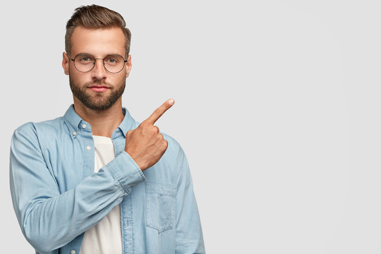Studio shot of attractive bearded man with thick beard and stylish haircut, points with fore finger at upper right corner, shows blank space for your advertising content or promotion. Look there!