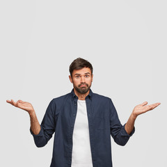 Vertical shot of hesitant puzzled employee shrugs shoulders with uncertainty, cannot make choice or understand something, dressed in fashionable shirt, stands against white wall with copy space