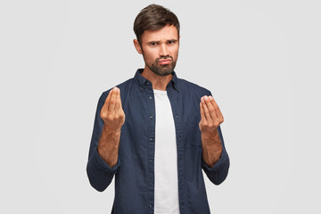 Handsome brunette man with thick dark beard makes money gesture, thinks about profitable business, demonstrartes symbol of currency and richness, tries to specify his mind. Body language concept