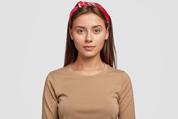 Portrait of pleasant looking European brunette girl wears stylish red headband, beige sweater,...