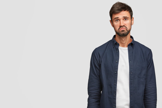 Sad uncertain displeased young man purses lips in discontent, raises eyebrows, dressed casually, isolated over white background with copy space for your advertisement or promotional content.