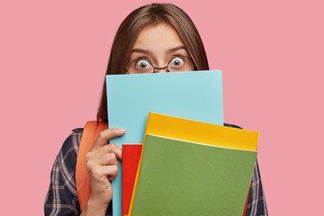 Back to school concept. Image of surprised emotive European woman hides behind books, has eyes...