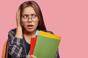 Emotive scared young woman keeps hand on head, looks desperately aside, opens mouth from...