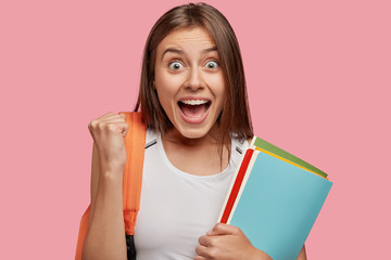 Overjoyed pupil girl with happy amazed expression, raises fists, rejoices successfully passed exam,...