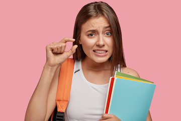 Puzzled European woman or pupil gestures with hand, shows something very little, demonstartes how good she knows material, poses against pink background with colourful folds. Youth, education concept