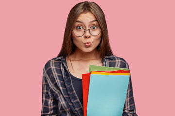 Attractive businesswoman holds business folders, makes grimace, dressed in checkered shirt, ready...