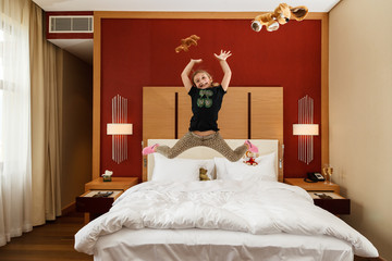 Young happy small Caucasian girl having fun jumping in the air with her toys above the bed in bedroom