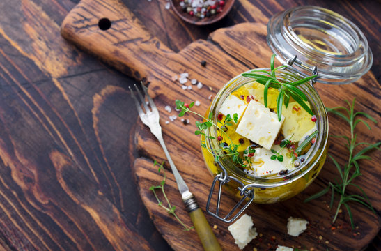 Feta cheese marinated in olive oil with fresh herbs in glass jar. Wooden background. Top view. Copy space.