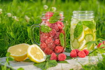 Fototapeta na wymiar Lemonade with raspberries and lemonade with lemon, cucumber and mint in a hipster jar for jam on a background of green grass. Concept: a picnic with refreshing drinks