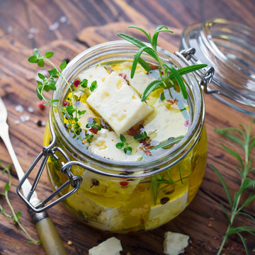 Feta cheese marinated in olive oil with fresh herbs in glass jar. Wooden background. Close up.