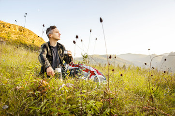 A young traveler sits on a hill in the background of a mountain and drinks tea from a thermos