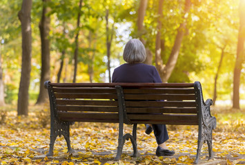 Lonely elderly woman sitting on a bench in autumn park.