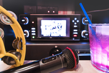 Black microphone in karaoke club, with remote controller, melon and strawberry soda drinks, yellow tambourine and screen for singing music on stage party. - obrazy, fototapety, plakaty