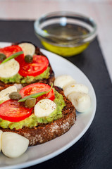 Sandwich toasts with tomatoes cherry, mozzarella, avocado, basil and olive oil. Side view on a dark stone dish.