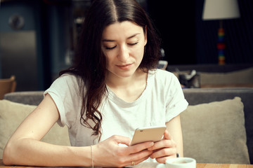 Young woman sitting in coffee shop at wooden table, drinking coffee and using smartphone. White cup...