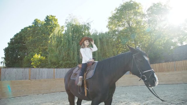Pretty little girl in hat riding a black mare on the arena. 4K