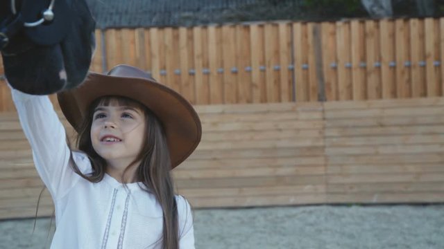 Cute little girl in hat comes and caresses black mare's head