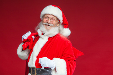 Christmas. Kind and tired Santa Claus in white gloves carries a red bag with gifts over his shoulder. Isolated on red background.