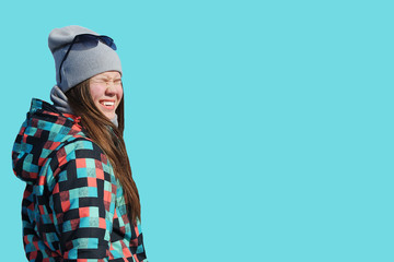 young girl in winter clothes and hat with glasses squints from sun and laughs isolated on blue background