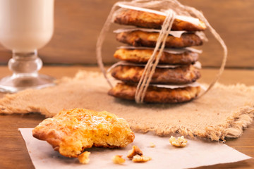 A stack of cookies, tied with a harsh thread, one cookie was broken and put on a paper napkin, a glass of milk, all on a wooden background.