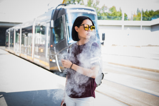 Stylish girl smoking an e-cigarette while standing near to road waiting for public transport