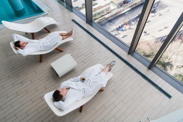 Two young man wearing bathrobe, lying on lounger in spa salon and talking to each other in front of a big window with a great city view. Spa concept