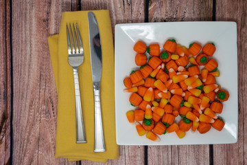 Plated view of candy corn and candy pumpkins with fork and knife on wooden table. Concept for Halloween candy for breakfast or dinner