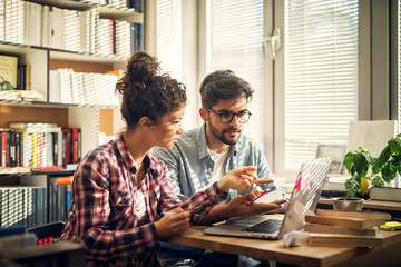 Two young modern students sitting in library and trying to solve some studying problems.