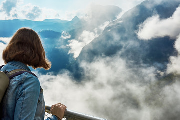 A young woman with a backpack standing by the railing over the mountains with clouds and mist looking down into the valley of Hallstatt. Austria in Europe