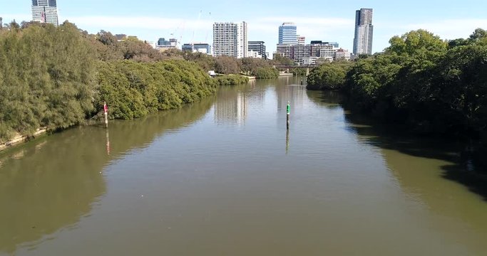 Low over calm waters of Parramatta river between tree covered shores towards bridge in front of Parramatta CBD towers and buildings.
