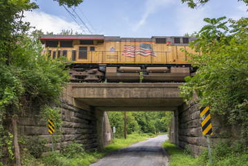 An orange train streaks across a concrete and stone bridge overpass and a lonely country gravel road.