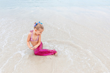 Little beautiful girl dressed in swimsuit as a mermaid sits on the seashore