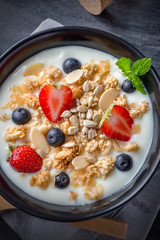 Vertical bowl of oat granola with yogurt, fresh strawberry, blueberries and nuts on black wooden table.