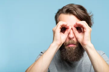 Foto op Canvas funny ludicrous joyful comic playful man pretending to look through binoculars made of hands. portrait of a young bearded guy on blue background. emotion facial expression concept © golubovy