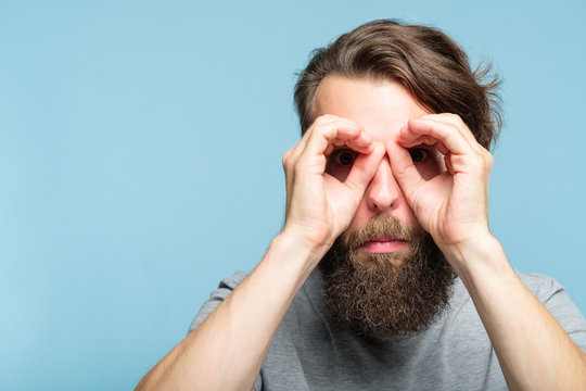 funny ludicrous joyful comic playful man pretending to look through binoculars made of hands. portrait of a young bearded guy on blue background. emotion facial expression concept