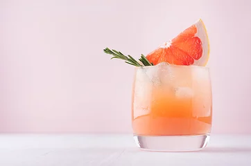 Wall murals Cocktail Cold fresh homemade grapefruit cocktail with ice, rosemary and pieces citrus on pastel pink background.