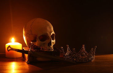 Human skull, old book, sword, crown and burning candle over old wooden table and dark background.