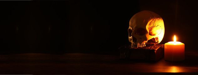 Human skull, old book and burning candle over old wooden table and dark background. - obrazy, fototapety, plakaty